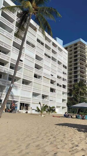 Ein mehrstöckiges Hotel mit Balkonen, gelegen an einem sandigen Strand mit Palmen.