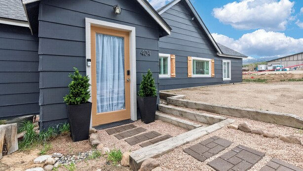 A modern two-story house with a dark exterior, a front door with a curtain, two potted plants, and a driveway leading to a garage. - 404 Doc Susie home Delightful and renovated home, walk to downtown, mt views (Fraser)