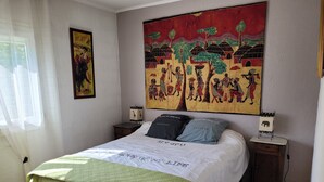 Una cama con colcha verde, una colcha blanca con texto, dos almohadas, una mesita de noche con un altavoz y un cuadro en la pared.