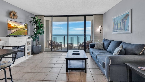 A living room with a sofa, coffee table, TV, and a balcony with a view of the ocean. - Ocean Forest Plaza 912 (Myrtle Beach)