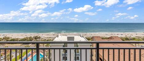 A view from a balcony overlooking a beach with a pool and a building with multiple floors and balconies.