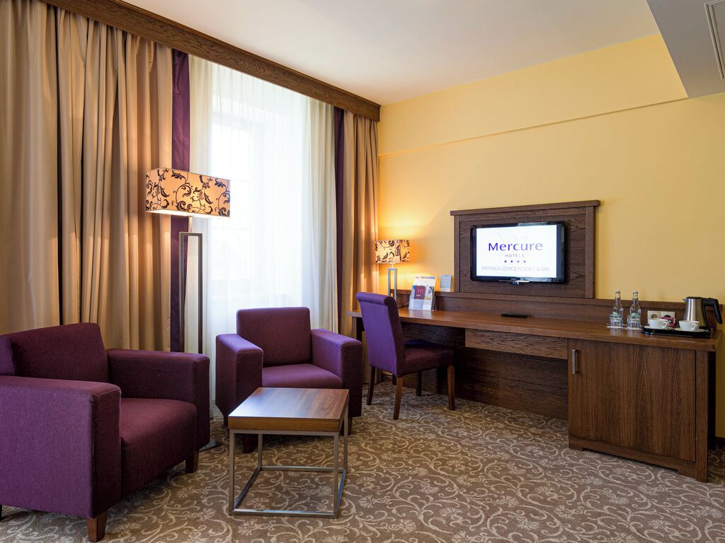 A hotel room with a desk, two purple armchairs, a sofa, a television, and a minibar.