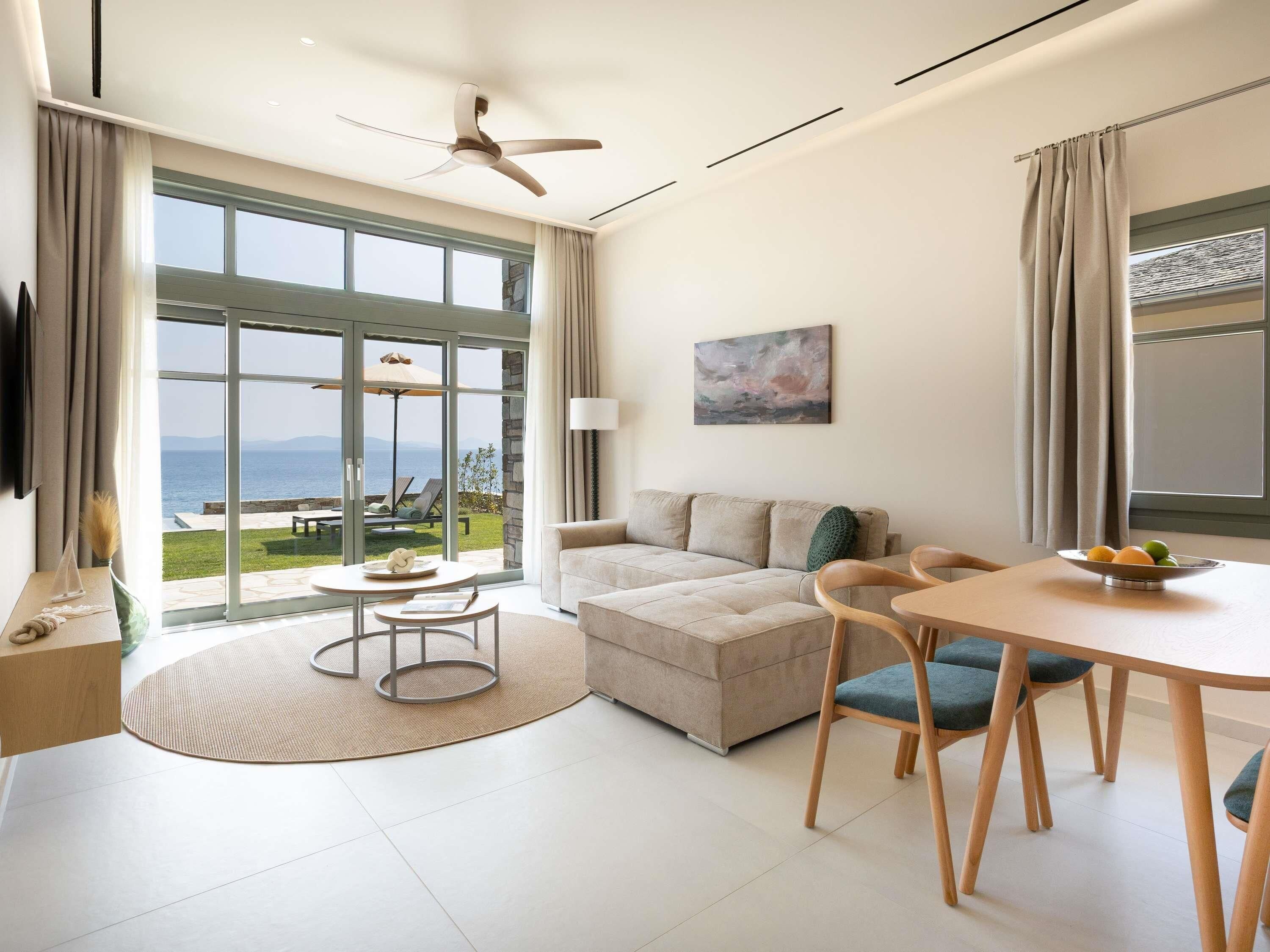 A modern living room with a sofa, dining table, chairs, and a view of the sea through large windows.
