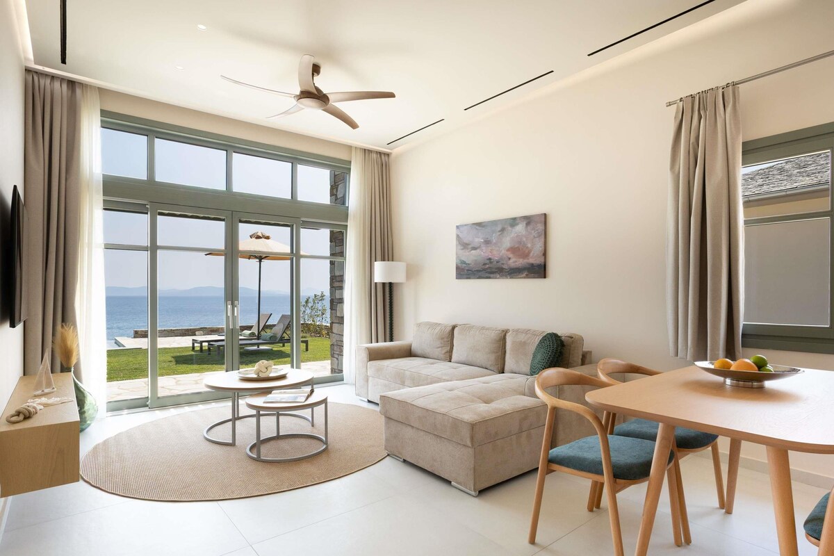 Ein modernes Wohnzimmer mit einer Couch, einem Esstisch, Stühlen und Blick auf das Meer durch große Fenster.