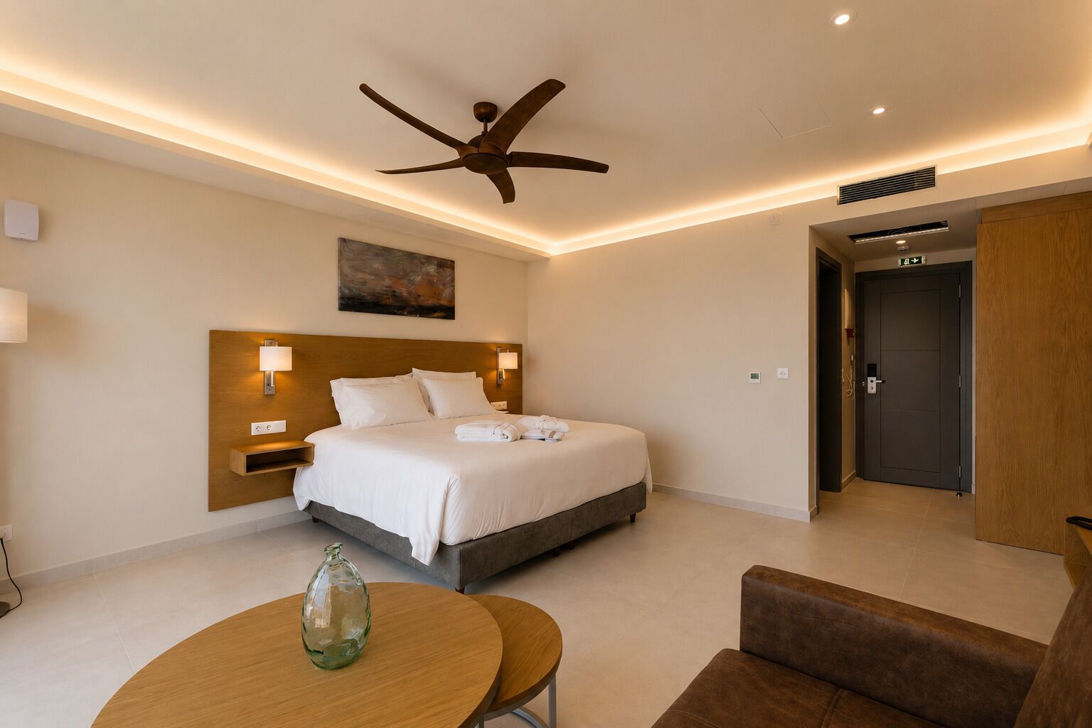 Quarto de hotel com cama, luminárias de cabeceira, ventilador de teto, um quadro, uma mesa redonda e uma cadeira.