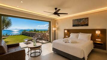 A hotel room with a bed, a desk, a chair, a table, a ceiling fan, and a view of the sea.