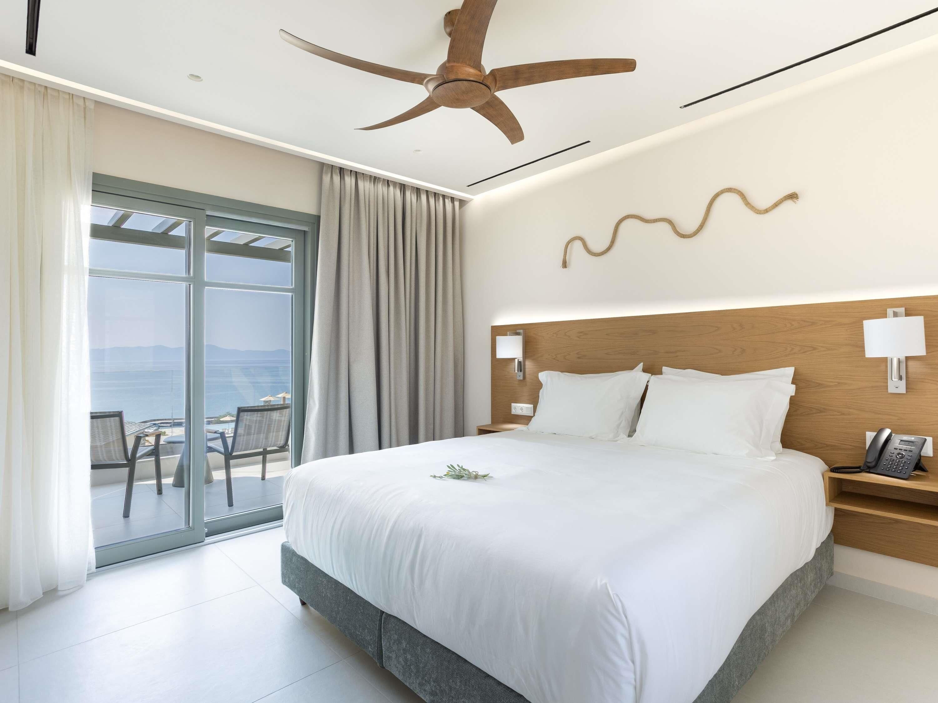 Una camera d'albergo con un letto, una ventola a soffitto, vista mare e un balcone con sedie.