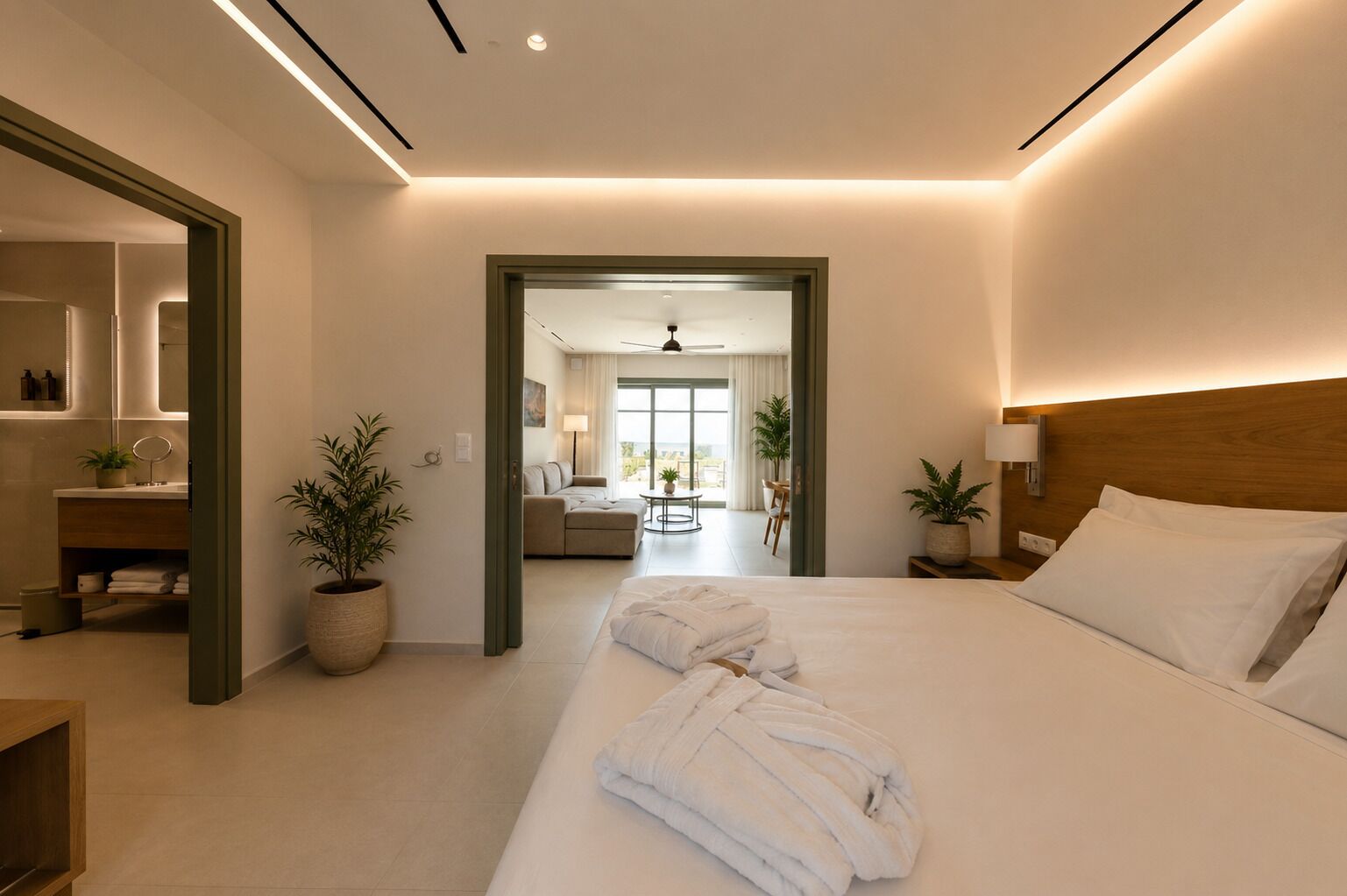 A modern hotel room with a large bed, a sitting area with a sofa and a table, a bathroom with a mirror and sink, and potted plants.