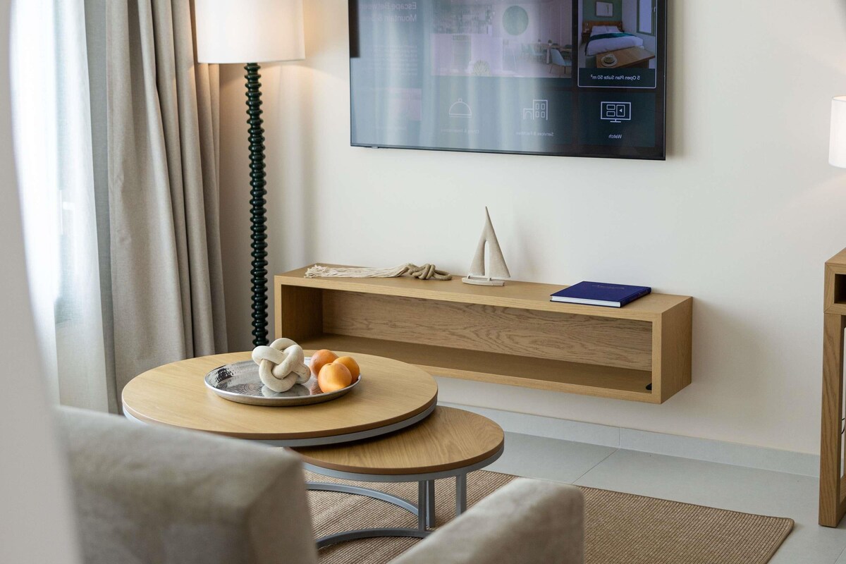 Ein modernes Hotelzimmer mit einem wandmontierten Flachbildfernseher, einem Holzkonsolentisch mit einer dekorativen Segeljacht, einem runden Holztisch mit einer Brotschale und einer Stehlampe.
