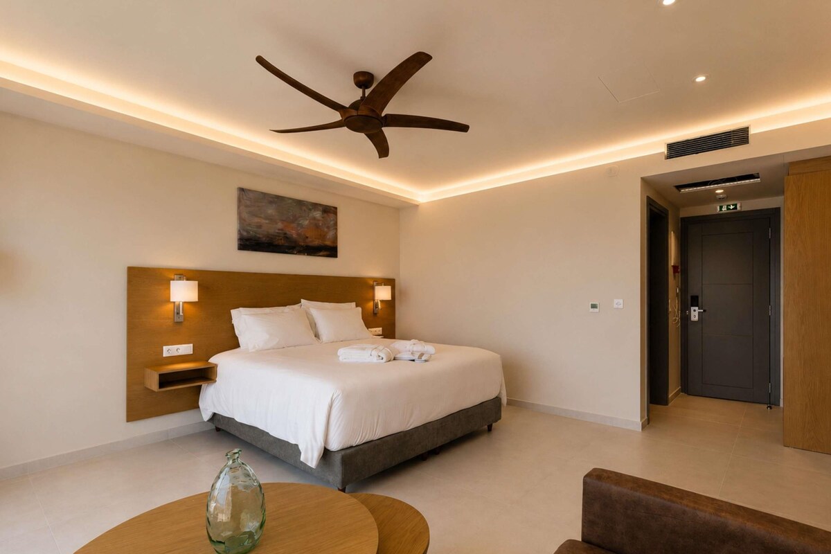 Ein Hotelzimmer mit Bett, Nachttischlampen, Deckenventilator, einem Gemälde, einer Tür und einem runden Tisch mit einer Glasflasche.