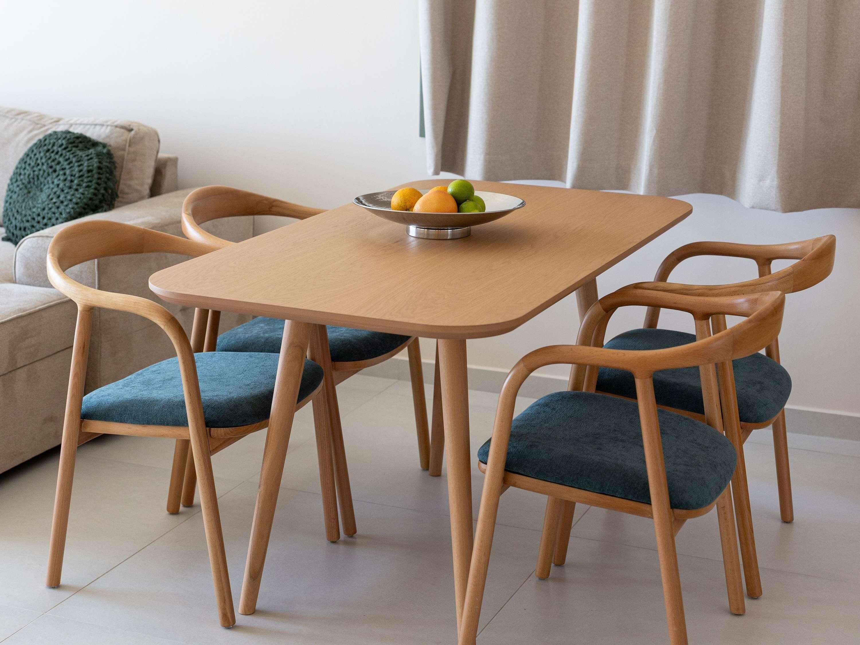 A dining area with a square wooden table, four wooden chairs with blue cushions, and a bowl of fruit on the table.