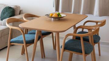 A dining area with a square wooden table, four wooden chairs with blue cushions, and a bowl of fruit on the table.