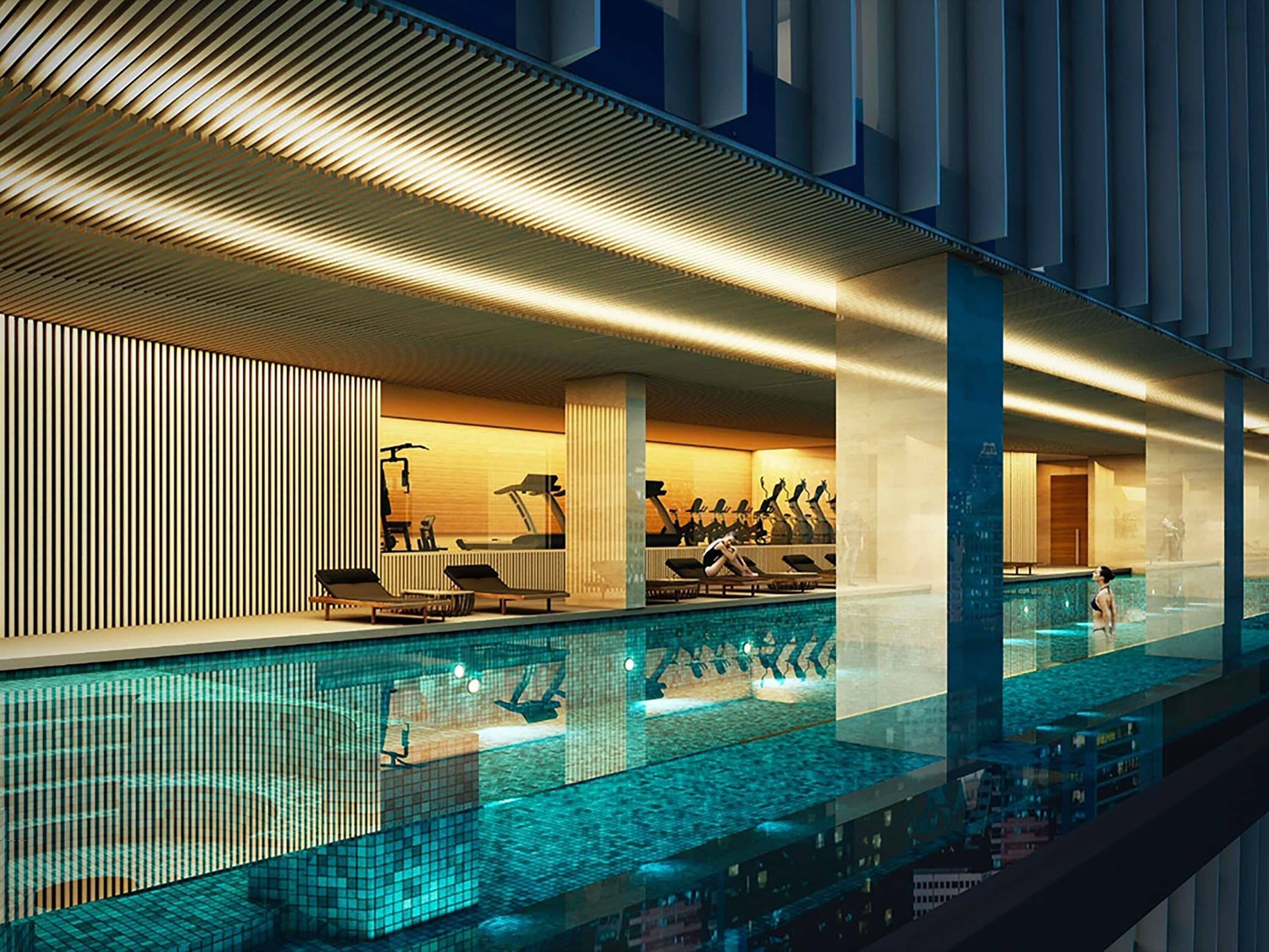 A modern indoor swimming pool with exercise equipment, a lounge area, and a view of the cityscape.