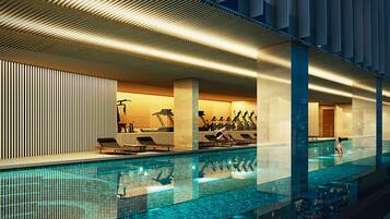 A modern indoor swimming pool with exercise equipment, a lounge area, and a view of the cityscape.