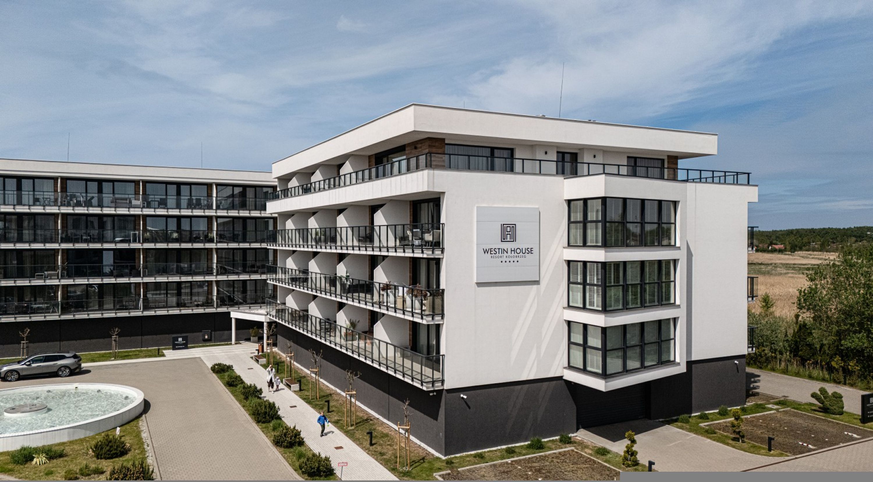 A modern building with multiple balconies, a sign reading 'WESTIN HOUSE RESORT FOUGGEREUSE,' and a parking area with a fountain.