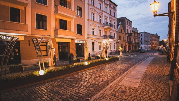Una strada acciottolata fiancheggiata da edifici a più piani, alcuni con insegne visibili, e un lampione.