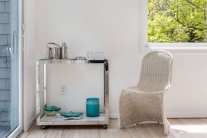 Ein Zimmer mit einem Korbstuhl, einem Chrom-Beistelltisch mit einer blauen Kerze und einem Fenster mit Blick auf Bäume.