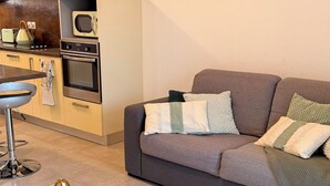 Een moderne woonkamer met een glazen salontafel, een grijze bank met decoratieve kussens, een keukenhoek met een magnetron en oven, en een roestvrijstalen afzuigkap.
