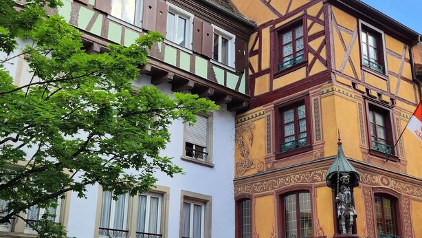 Ein traditionelles europäisches Gebäude mit einer Café-Terrasse, Sitzgelegenheiten im Freien und einer Flagge.