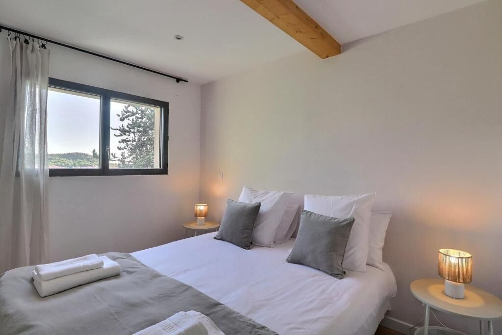 A bedroom with a bed, two bedside lamps, a window with curtains, and a view of greenery outside.