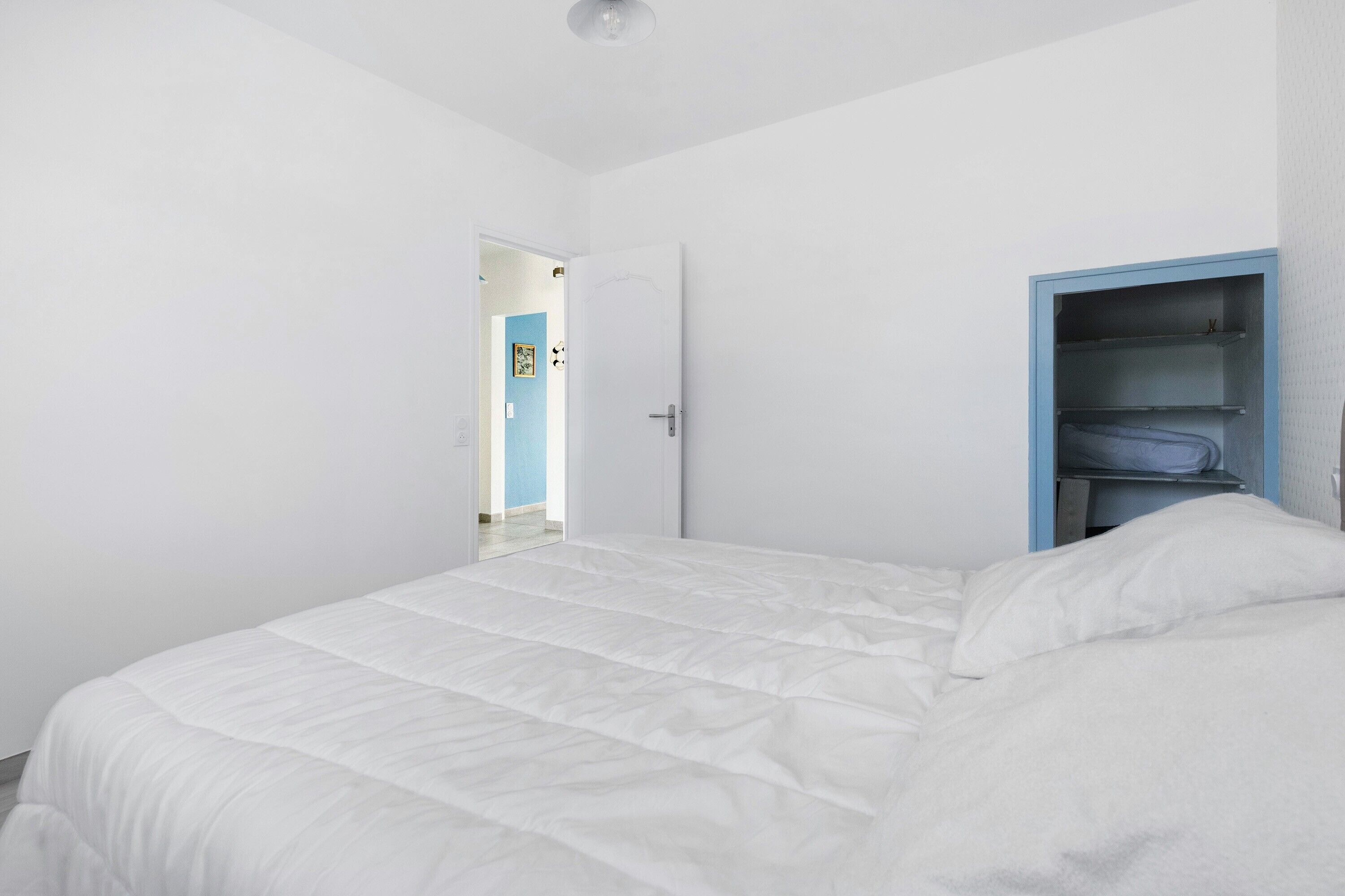 Un dormitorio con una cama blanca, un armario azul y una puerta que da a otra habitación.