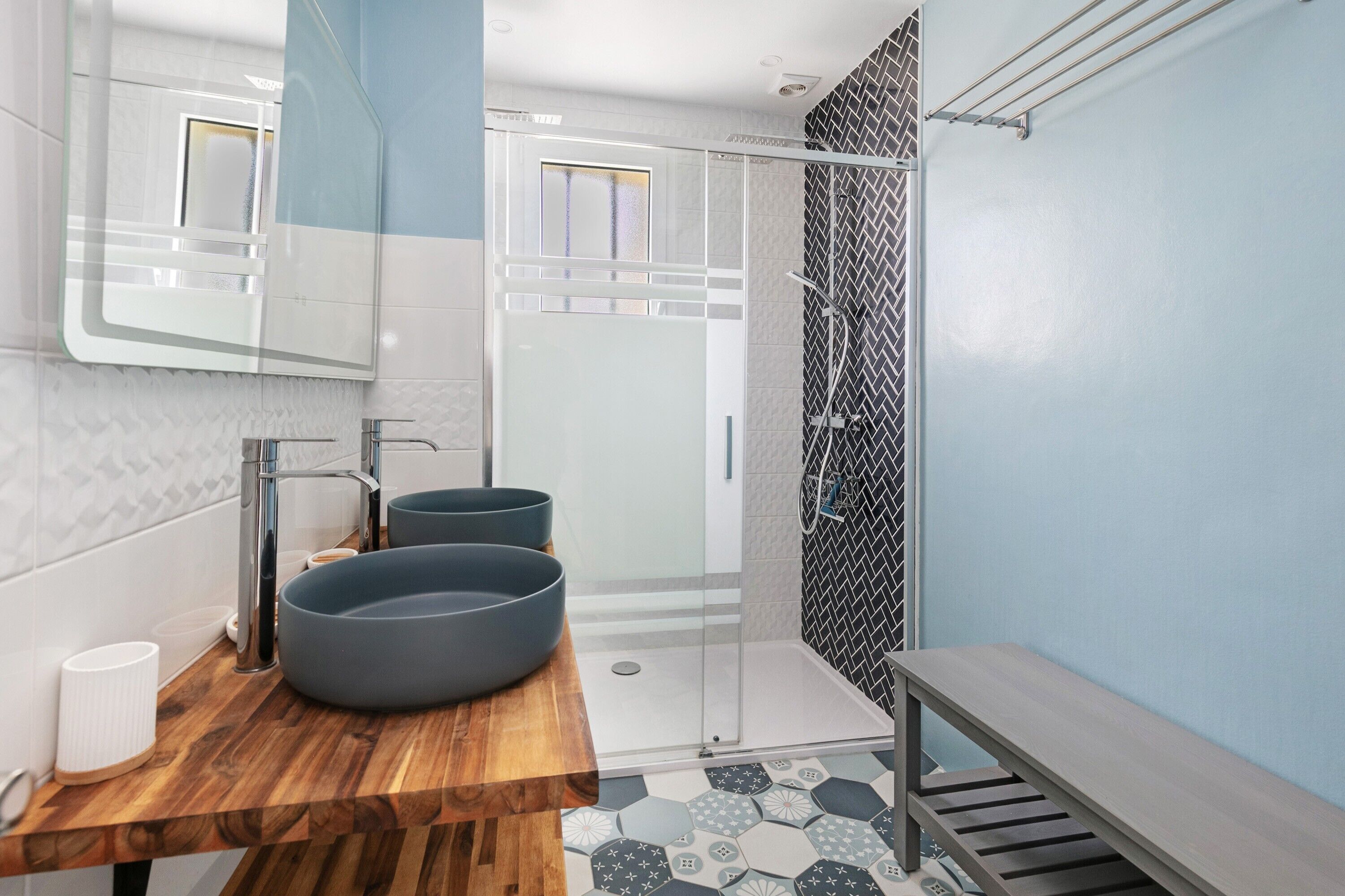 Un baño moderno con tocador de madera, dos lavamanos grandes, cabina de ducha de vidrio y pared con diseño.