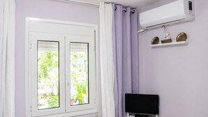Una stanza con una finestra, condizionatore, televisore su un mobile e scaffale con oggetti decorativi.