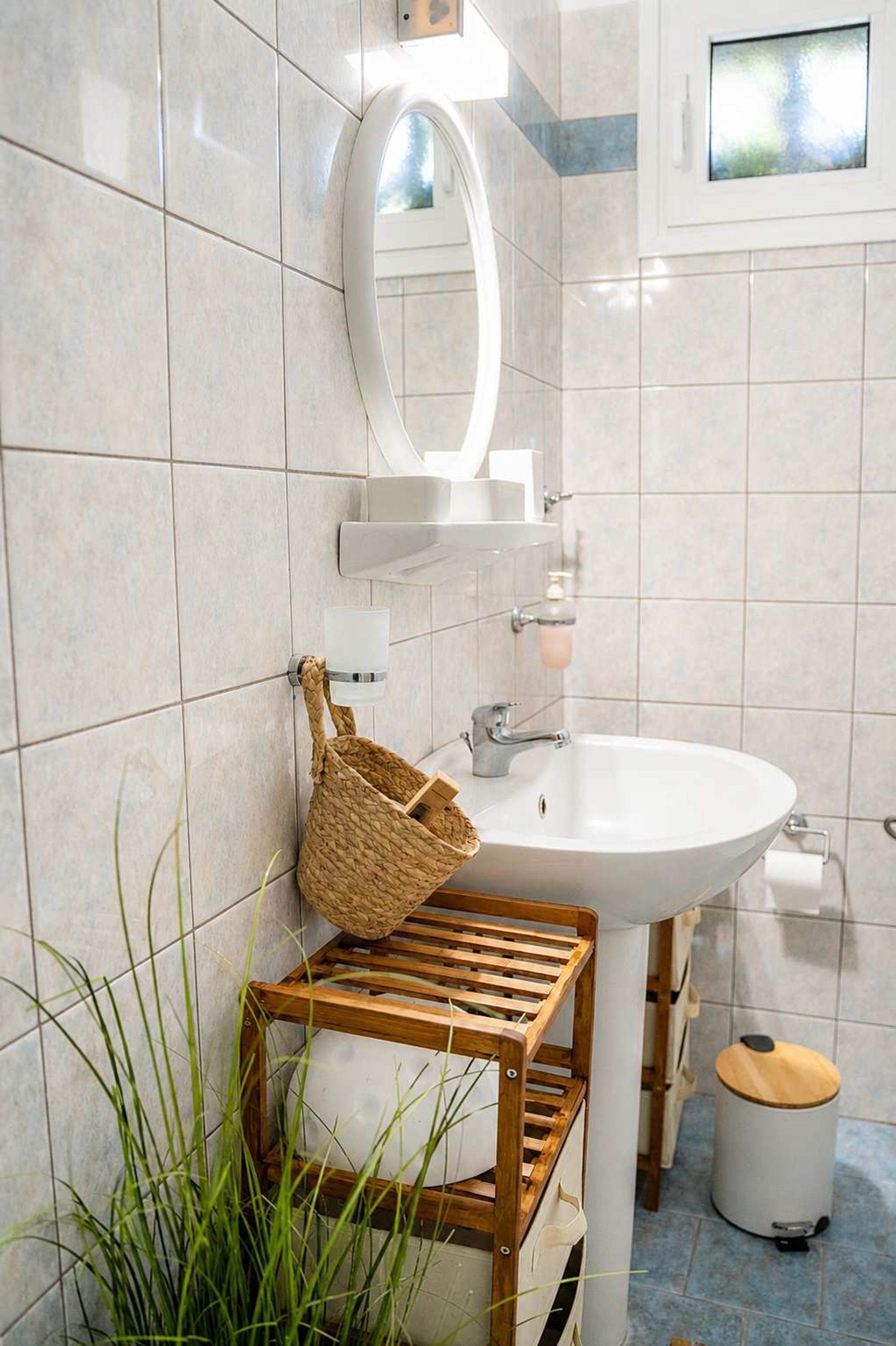 A bathroom with a white sink, a mirror, a towel rack, a trash bin, and a small window.