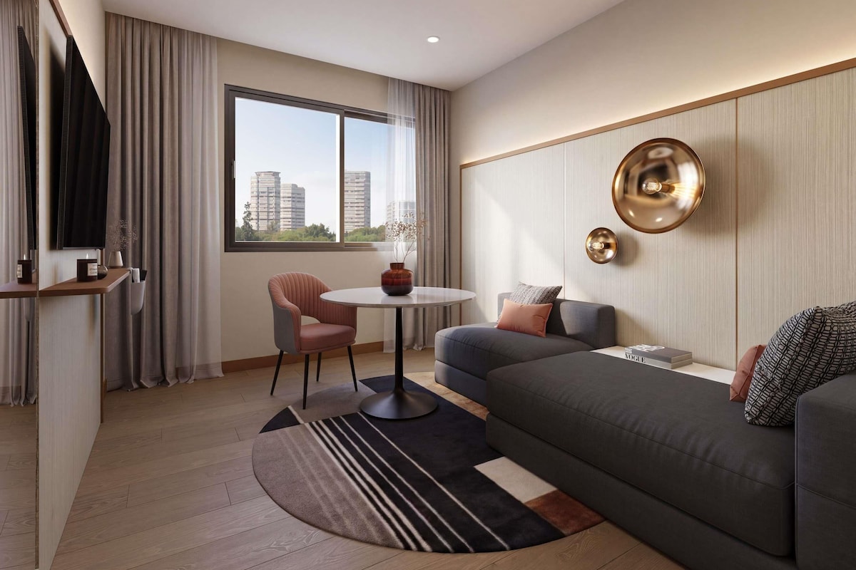 Habitación de hotel moderna con sofá, una mesita redonda, televisión y un ventanal con vistas a la ciudad.