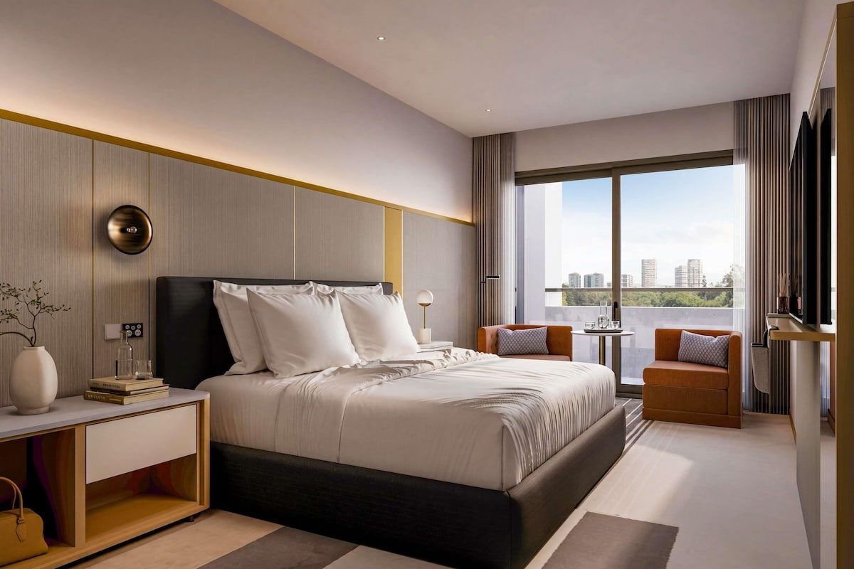 Un dormitorio de hotel moderno con una cama grande, mesitas de noche, un escritorio, una silla y vistas a la ciudad.