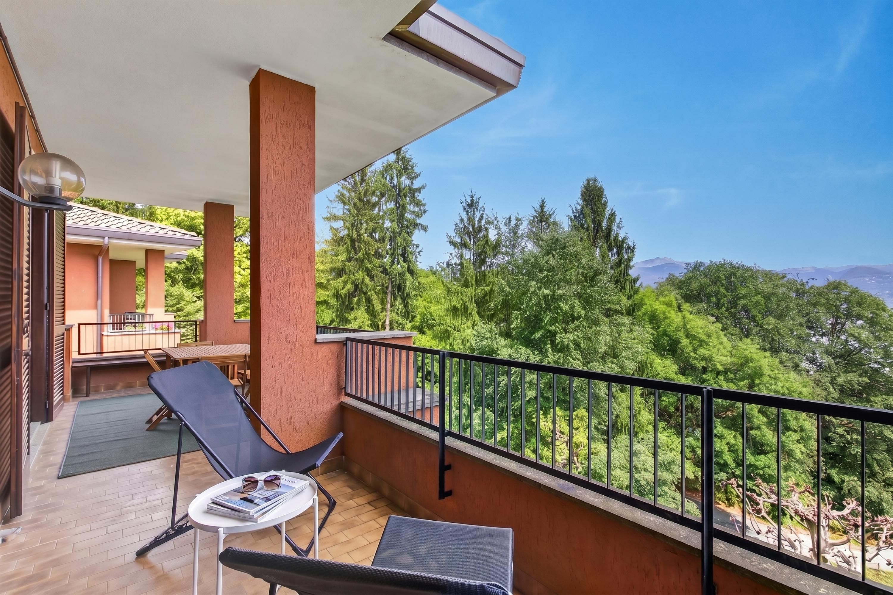 Un balcone con tavolo e sedie, vista su alberi e montagne, e un edificio con tetto in tegole.