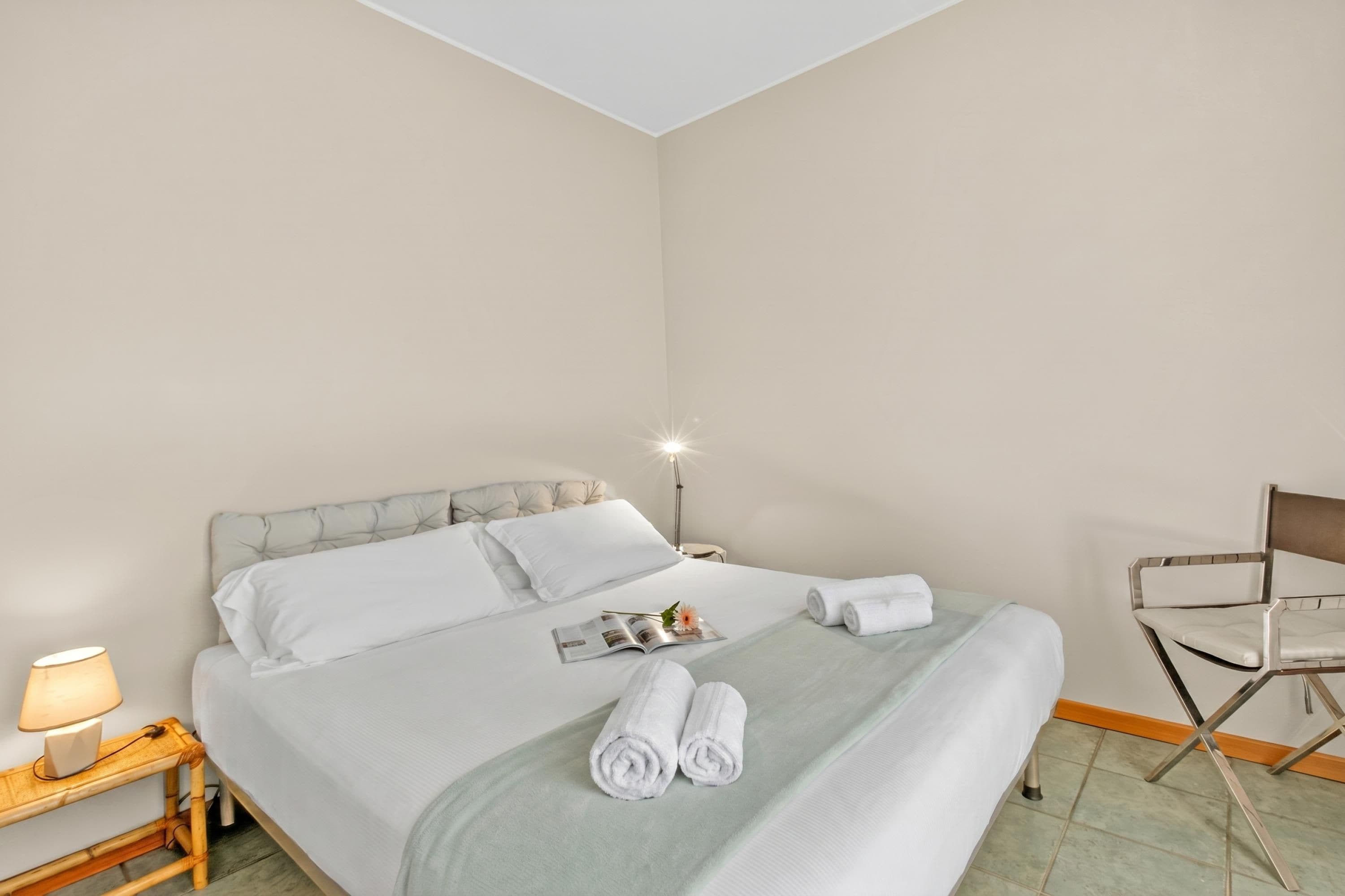 A neatly made bed with white linens, a lamp on a bedside table, and a chair in a hotel room.
