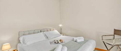 A neatly made bed with white linens, a lamp on a bedside table, and a chair in a hotel room.