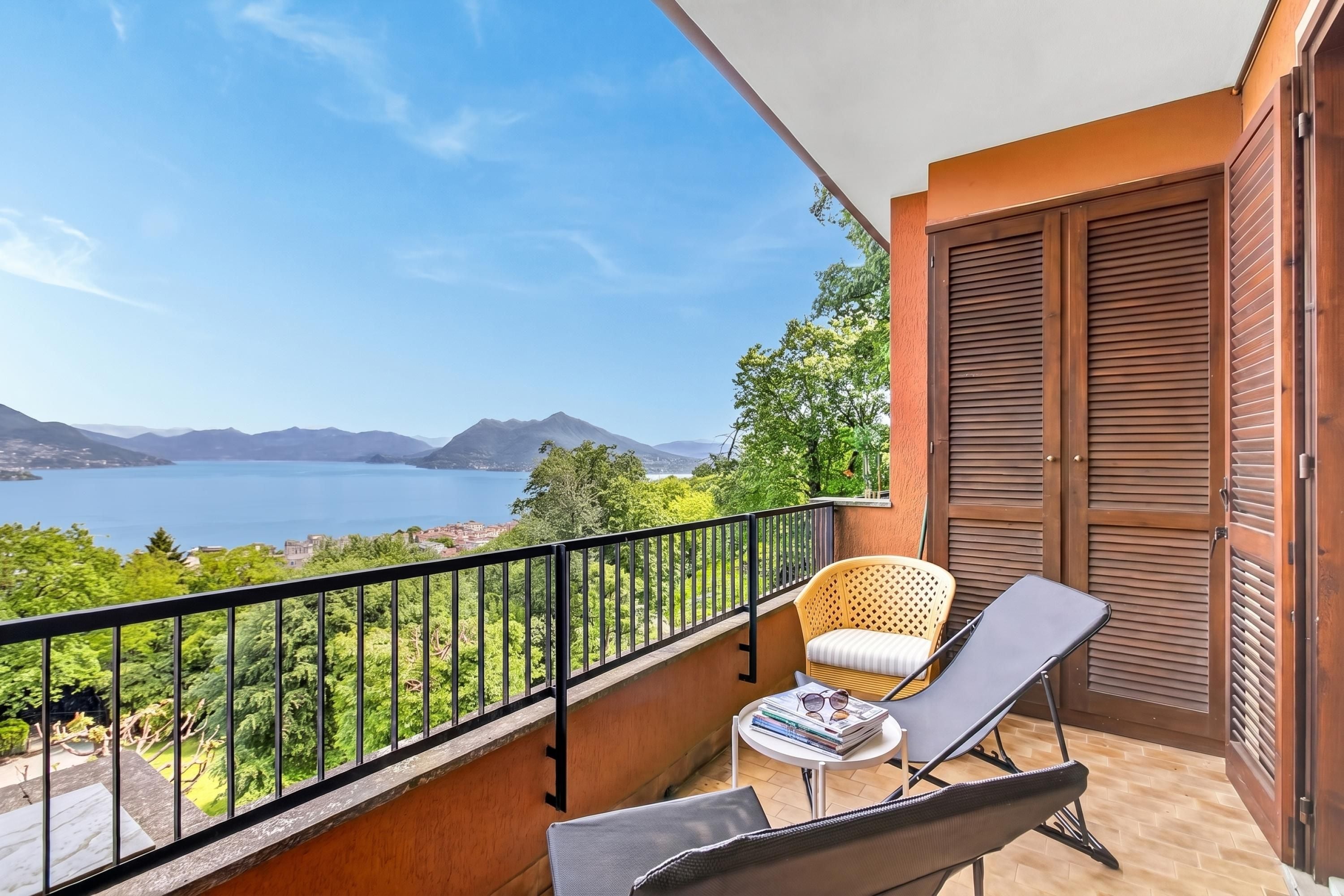 Ein Balkon mit Blick auf einen See und Berge, ein Stuhl, ein Tisch mit Büchern und Gläsern sowie eine Holztür.