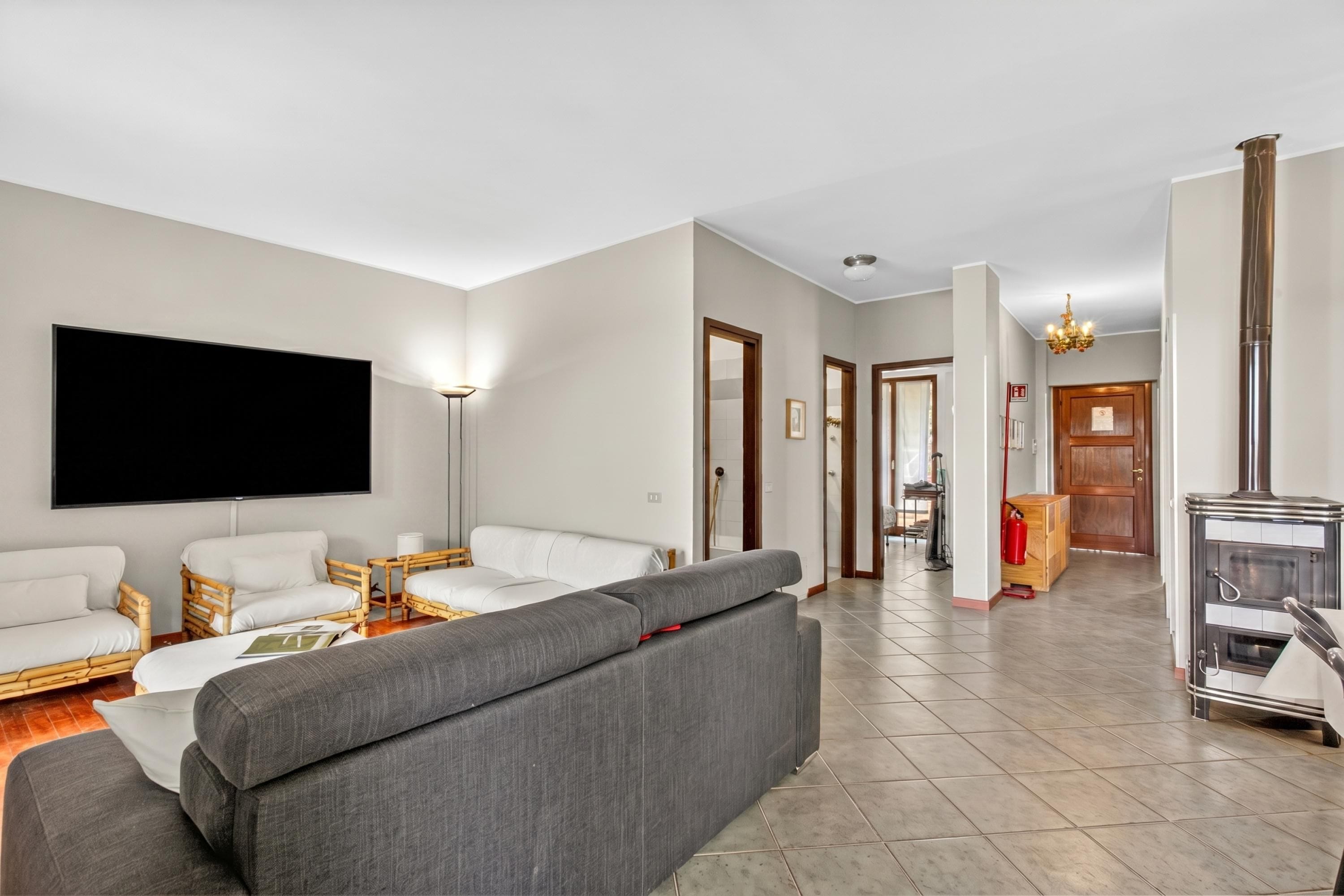 A living room with a large flat-screen TV, a grey sofa, white armchairs, and a wooden coffee table.
