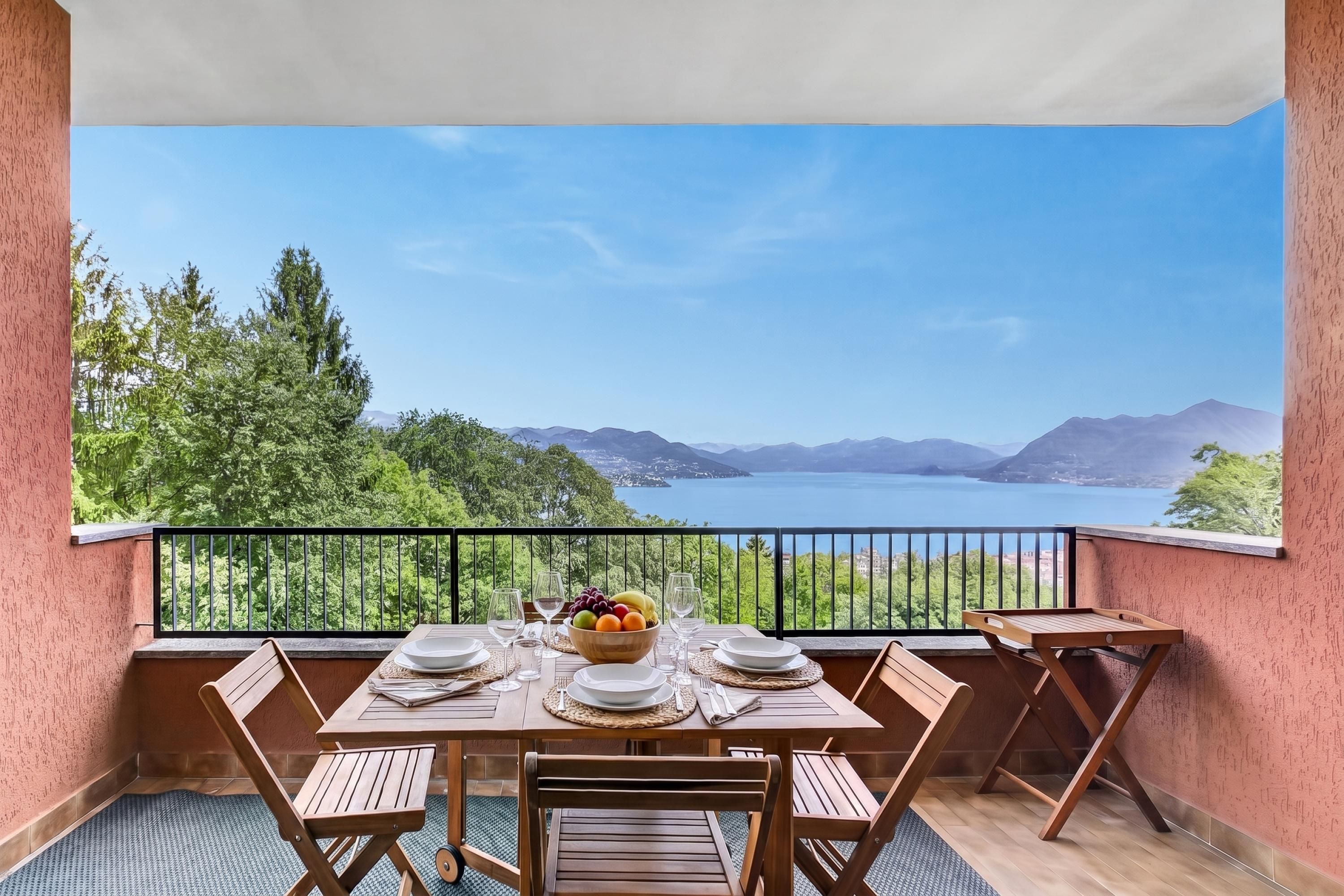 Un balcón con una mesa lista para dos personas, un frutero y vistas a un lago y montañas.