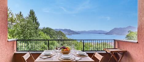 Un balcón con una mesa lista para dos personas, un frutero y vistas a un lago y montañas.
