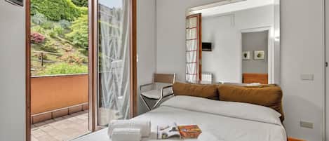 A hotel room with a bed, a chair, a small table, and a sliding glass door leading to a balcony with a garden view.