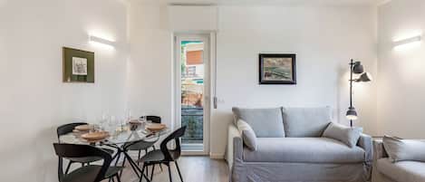 A modern living room with a grey sofa, a dining table with chairs, and a door leading to an outdoor area.