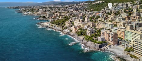 A coastal cityscape with buildings along the waterfront, a clear sky, and a mountainous backdrop.