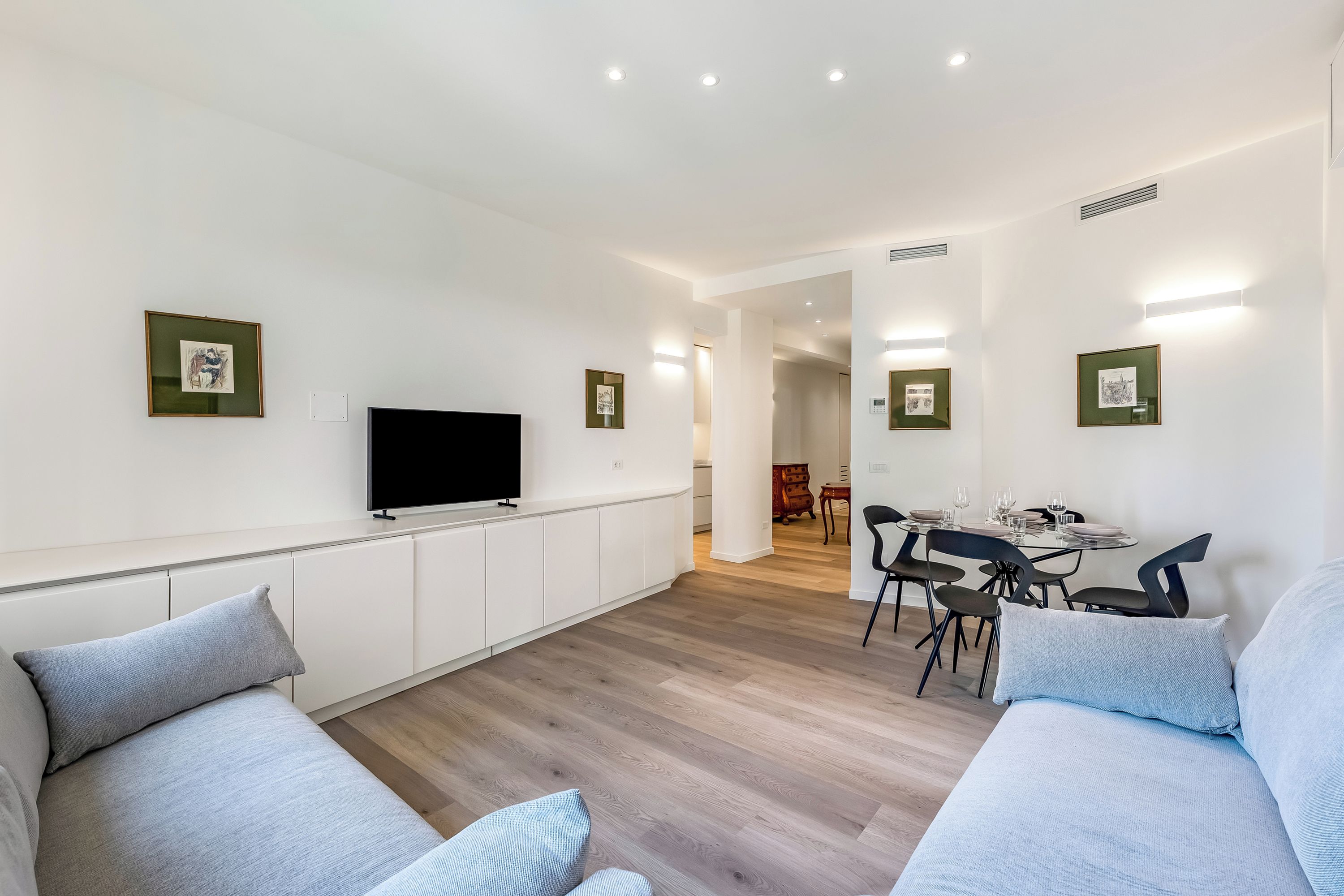 Una sala de estar moderna con un televisor de pantalla plana, sofás de gris claro, un comedor con mesa y sillas, y cuadros en las paredes.