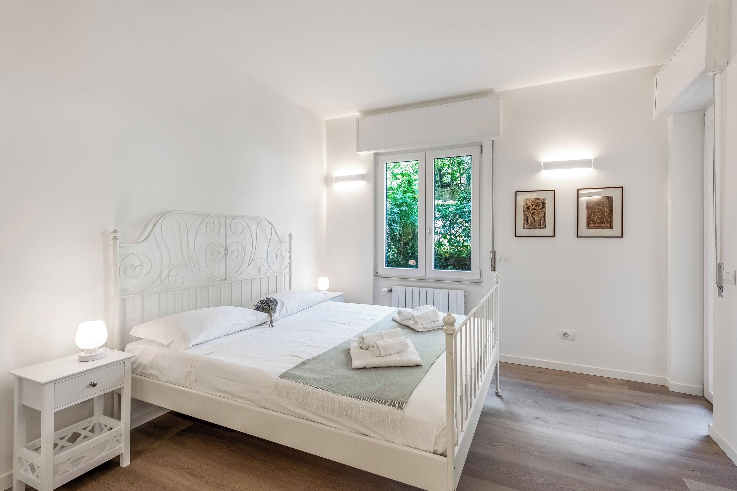 A white bed with a headboard, a bedside table with a lamp, a window with a view of greenery, and two framed pictures on the wall.