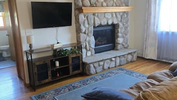 Un salon avec une cheminée en pierre, un téléviseur fixé au mur, un banc en bois et un tapis à motifs.