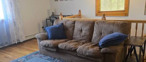 A living room with a brown leather sofa, a blue rug, a wooden dining table, and a ceiling fan.
