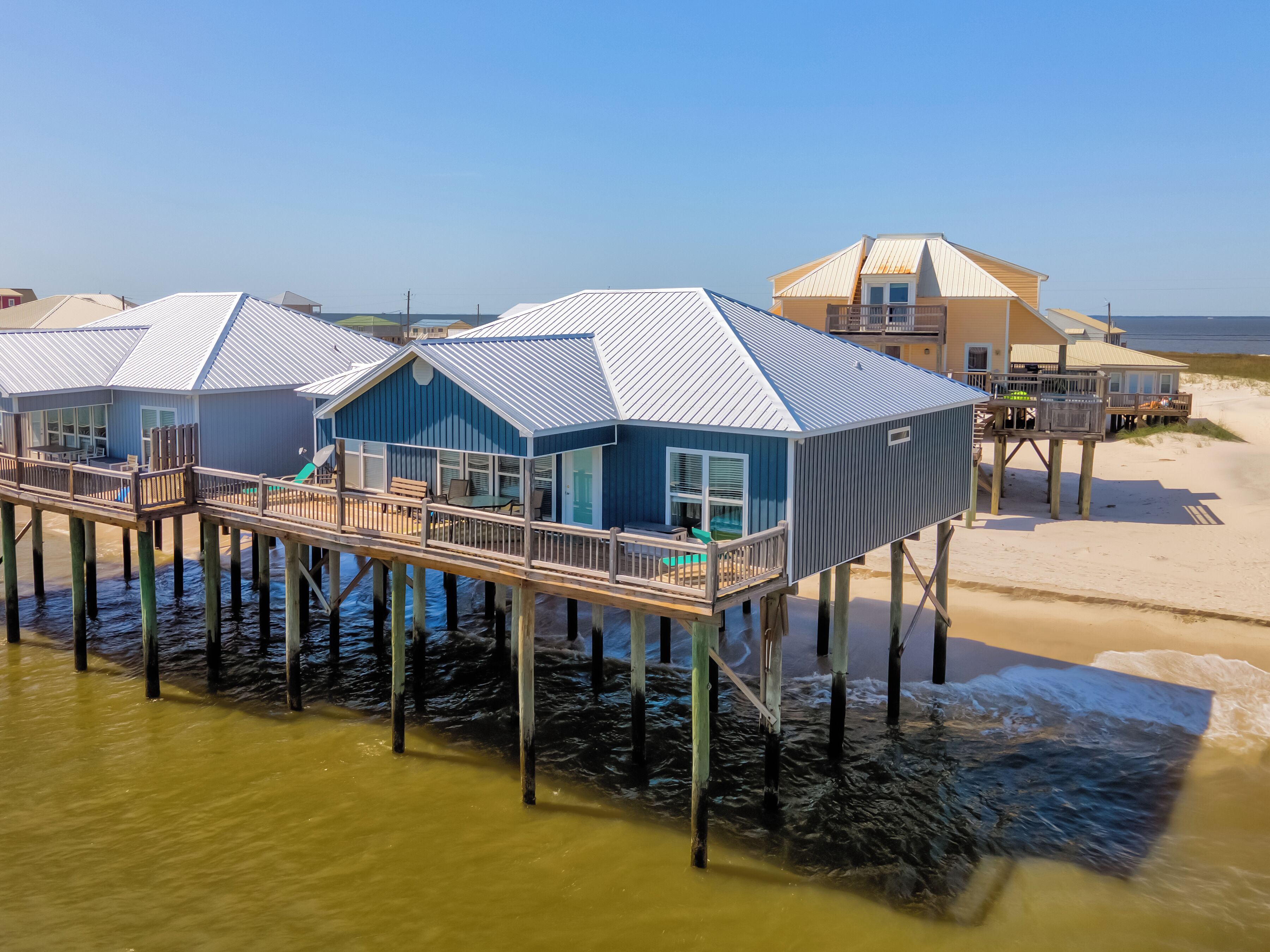 Une maison bleue avec un balcon sur pilotis au-dessus de l’eau, une terrasse et une plage en arrière-plan.