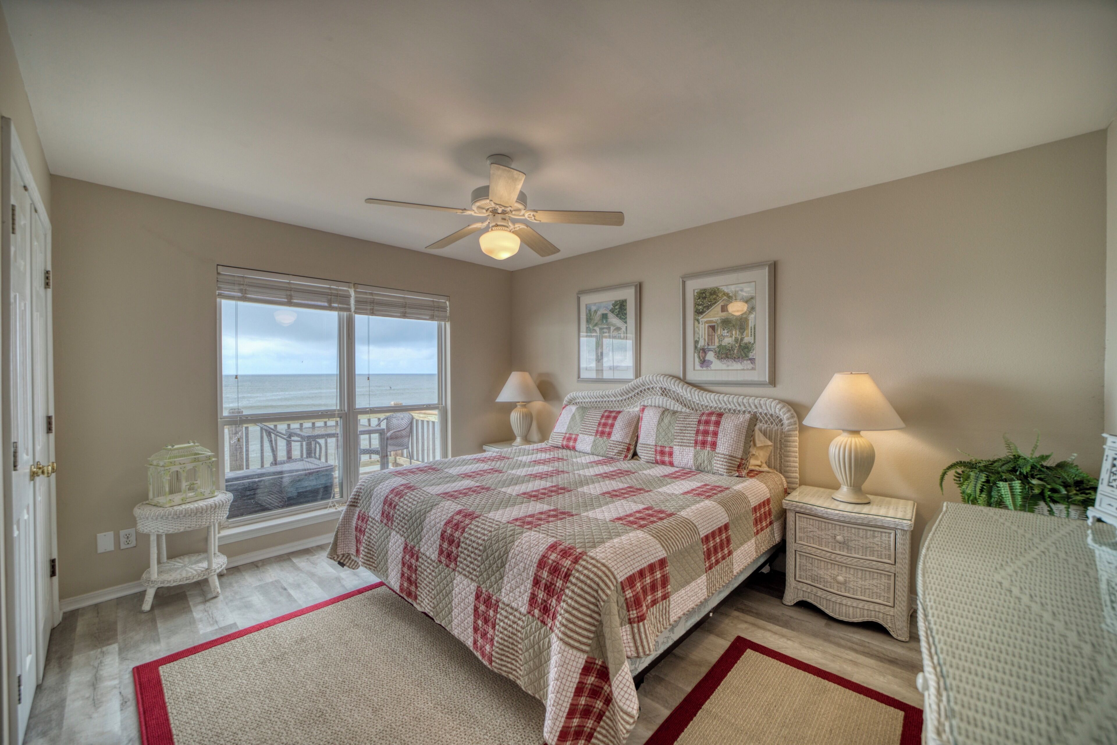 A bedroom with a large window, a ceiling fan, a bed with a checkered bedspread, two bedside tables with lamps, and a small white chair.