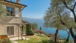 Ein Steinhaus mit Balkon, Terrasse und Blick auf einen See und Berge.