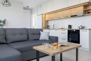 A modern kitchen with white cabinets, a built-in oven, and a dining area with a grey sofa and a wooden table. - Spacious Apartment with a Balcony | Parking (Lublin)
