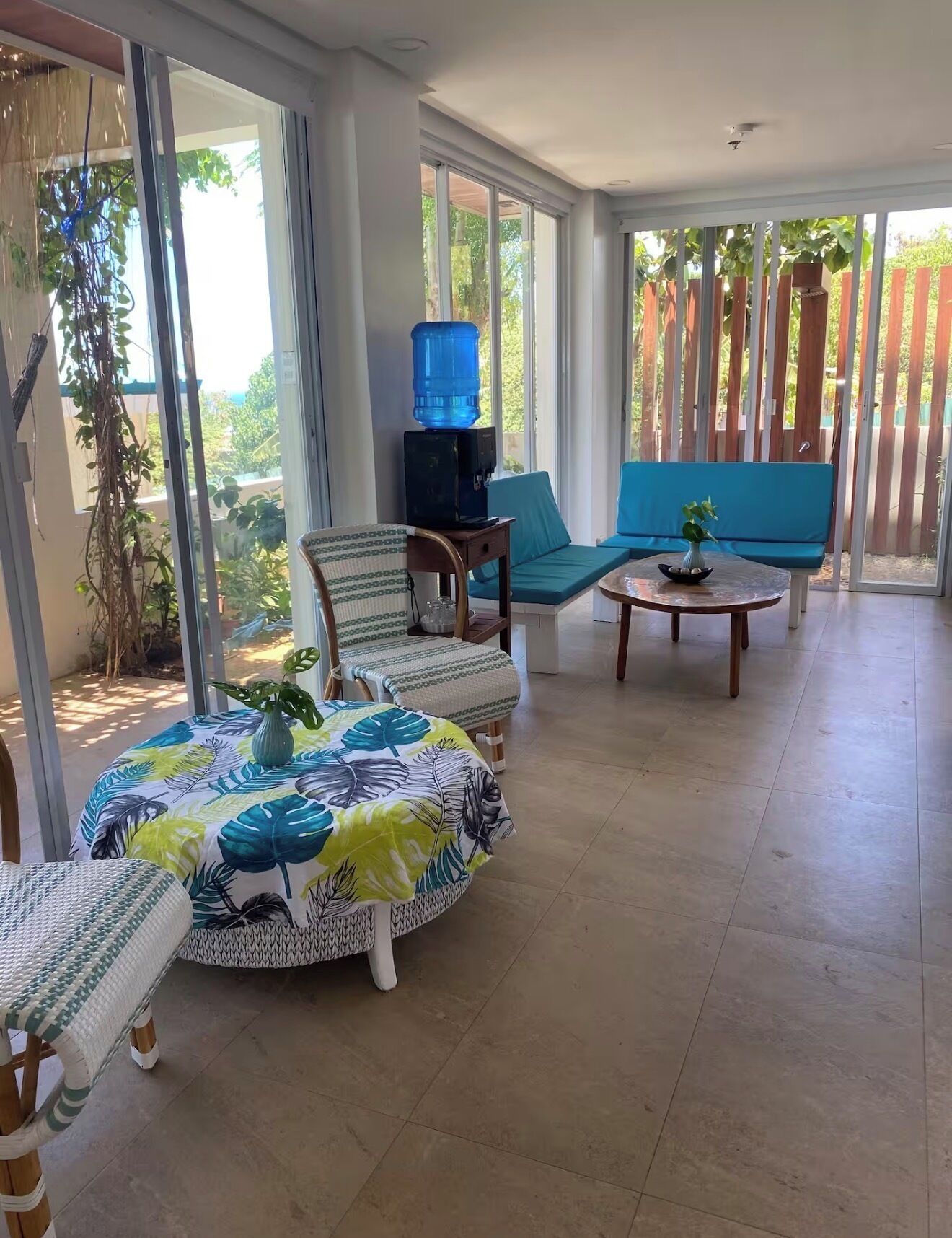 Une terrasse ensoleillée avec une table ronde, deux chaises, un canapé et un distributeur d’eau.