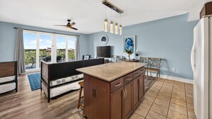Eine moderne Küche mit einer zentralen Kochinsel, einem Essbereich mit Stühlen, einem Deckenventilator und einem großen Fenster mit Vorhängen.